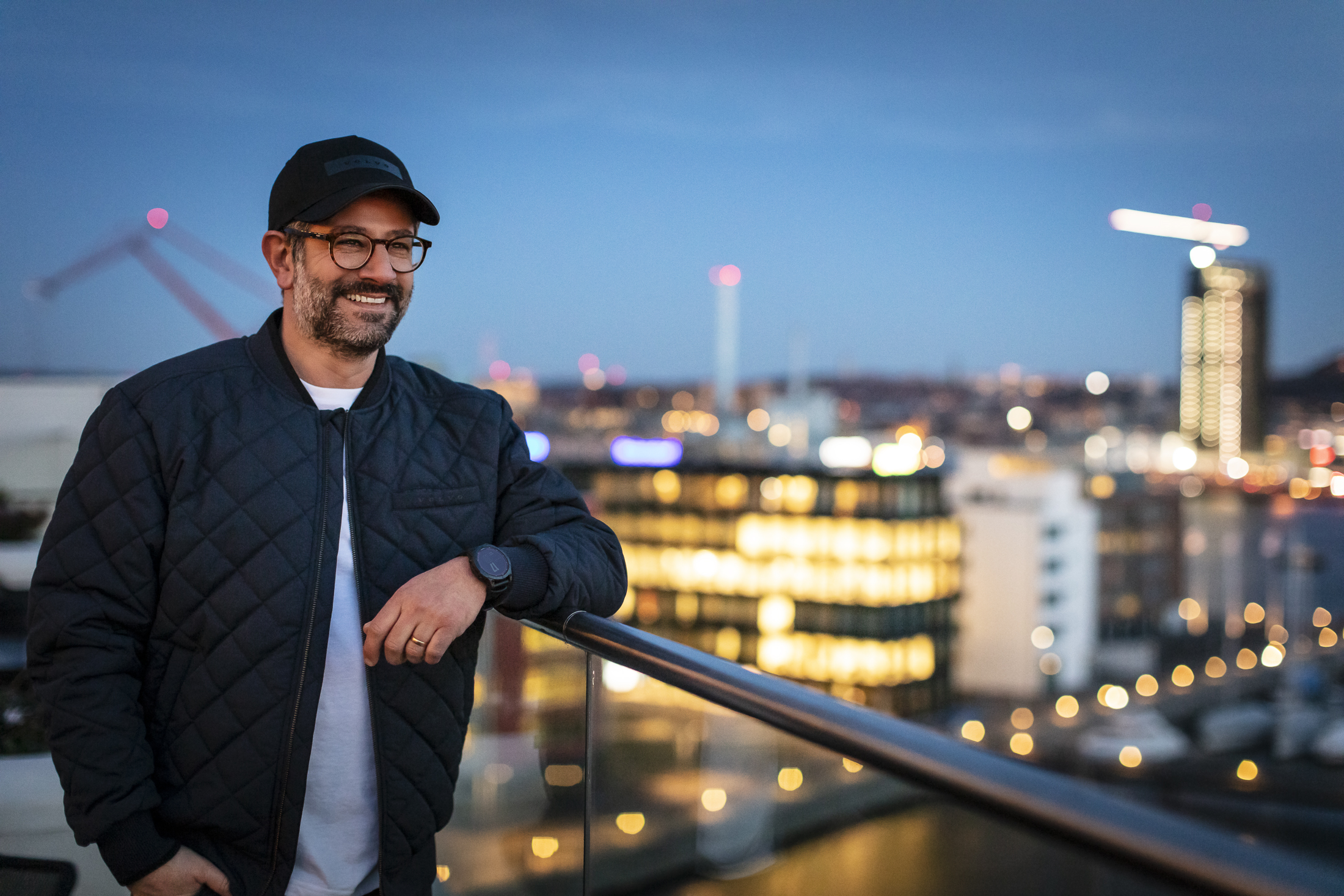 Man wearing Volvo navy jacket and black cap on a roof with city view